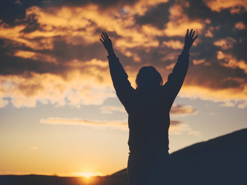 Photo of a person outside facing a stormy sky with hands raised.